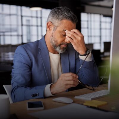 Mature businessman holding his head in stress, sitting at a desk with computer and documents.