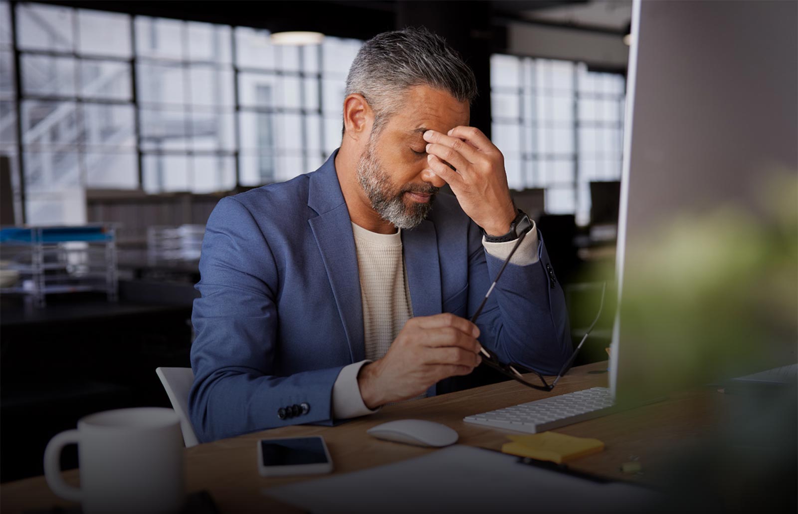 Mature businessman holding his head in stress, sitting at a desk with computer and documents.
